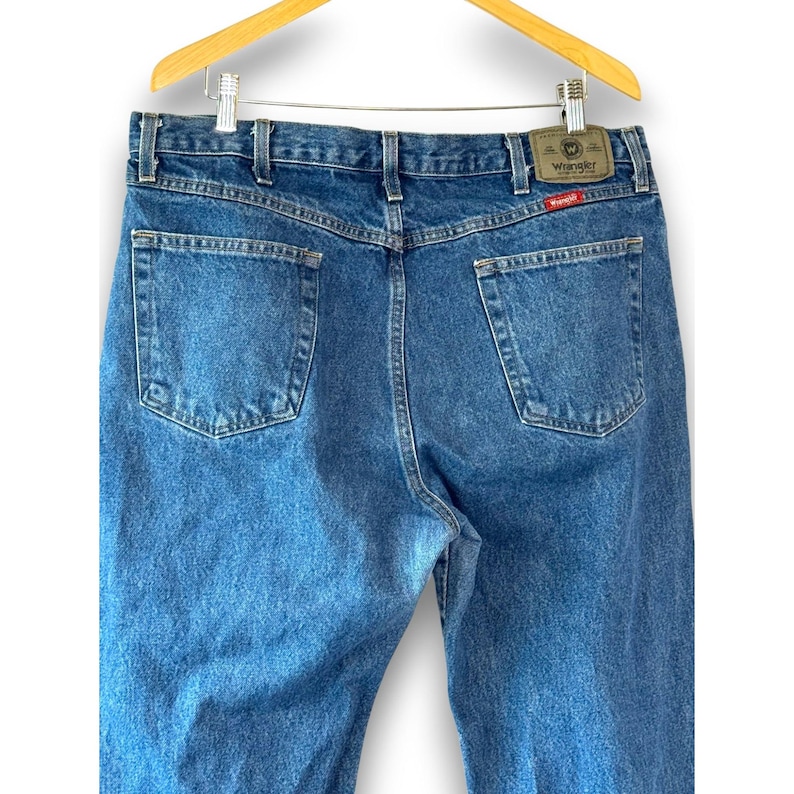 May include: Blue denim jeans with a classic five-pocket design, hanging from a wooden hanger. The jeans feature a straight leg cut and a Wrangler brand patch on the back. The wash is a medium blue.