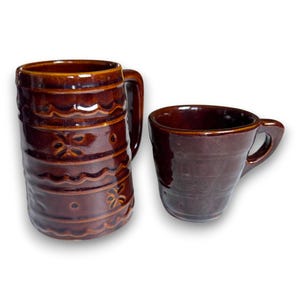 May include: Two brown ceramic mugs. One is a tall, cylindrical beer stein with a handle and decorative floral and wave patterns. The other is a smaller, rounded mug with a handle. Both mugs have a glossy finish.