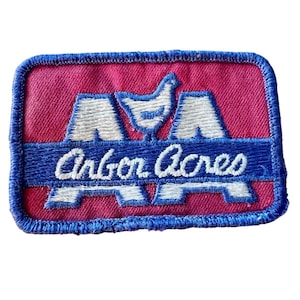 May include: A rectangular embroidered patch with a magenta background and a blue border. It features a white stylized chicken icon above the text Arbor Acres in white script, set against a blue horizontal bar, with white letter A shapes.