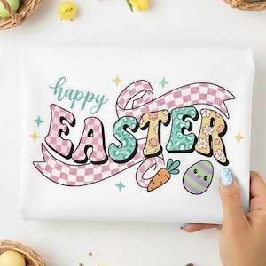 May include: White t-shirt with a retro-style "Happy Easter" design. The text is in pink, teal, and yellow, with a checkered ribbon and Easter egg and carrot illustrations. The design is surrounded by pastel stars.