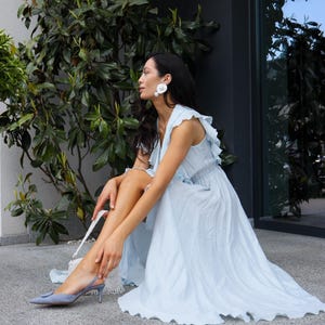 May include: A light blue sleeveless maxi dress with ruffled shoulder details and a textured fabric finish. The dress is paired with light blue pointed-toe slingback heels featuring a circular buckle detail and large white circular stud earrings.