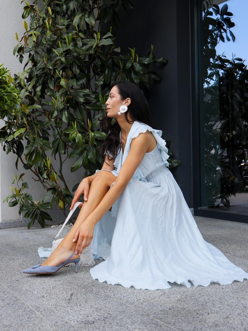 May include: A light blue sleeveless maxi dress with ruffled shoulder details and a textured fabric finish. The dress is paired with light blue pointed-toe slingback heels featuring a circular buckle detail and large white circular stud earrings.