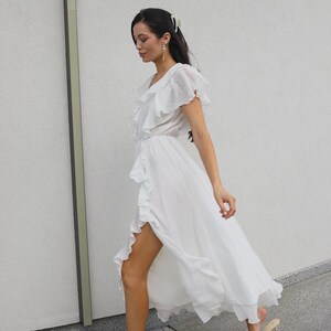 May include: A white midi dress featuring a V-neckline, short flutter sleeves, and a cascading ruffle detail that runs along the front opening. The lightweight fabric creates a flowing silhouette with a high side slit, paired with beige heeled sandals.