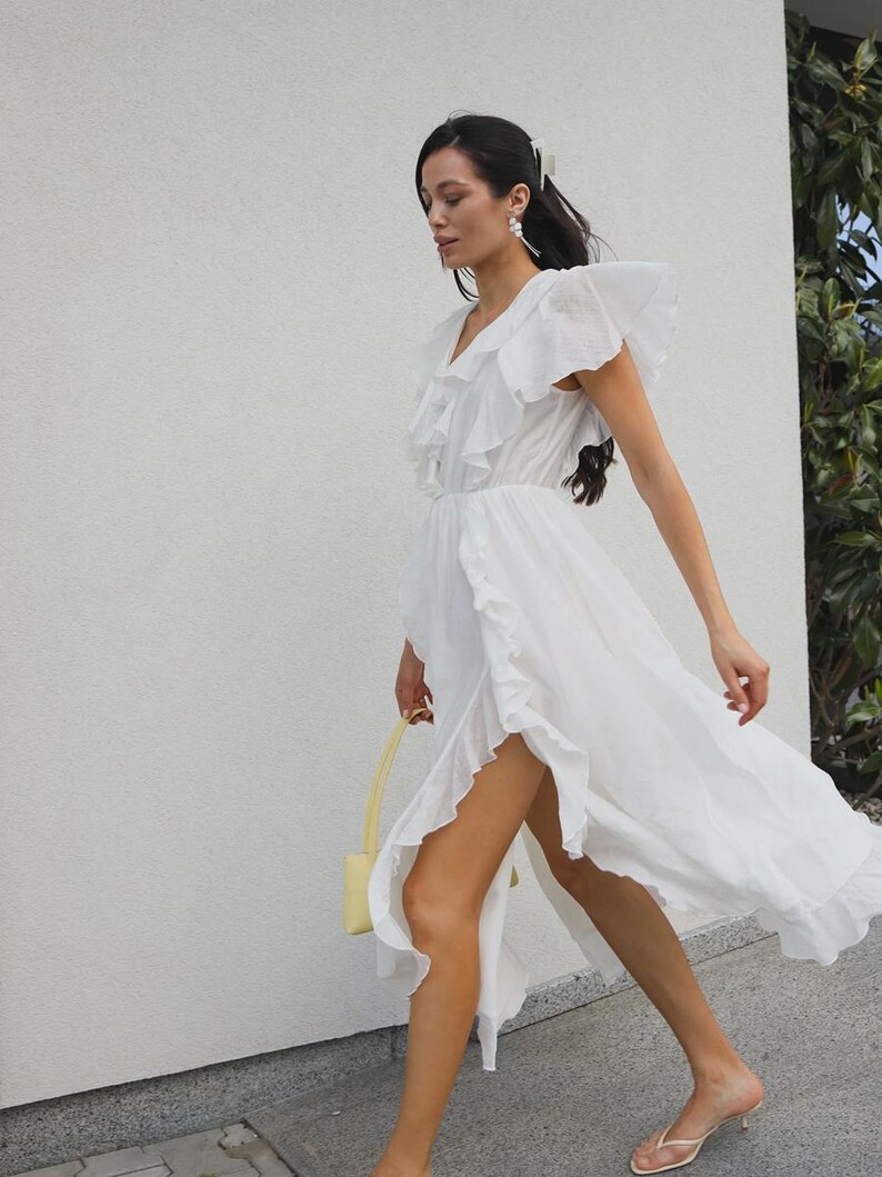 May include: A white midi dress featuring a V-neckline, short flutter sleeves, and cascading ruffles along the bodice and the high-low hemline. The lightweight fabric has a textured finish, and the skirt includes a side slit detail.