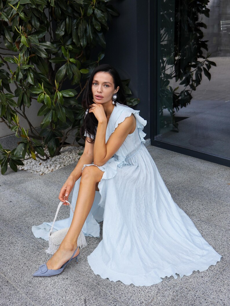 May include: A light blue sleeveless maxi dress featuring a textured fabric, a V-neckline, and ruffled shoulder details. The dress has a flowing skirt with a high side slit. It is paired with a small white beaded handbag and light blue slingback heels.