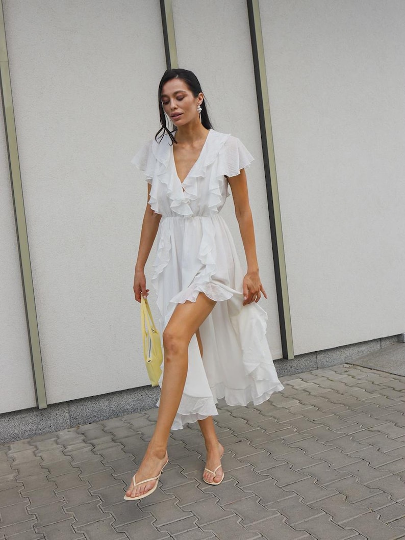 May include: A white midi dress featuring a deep V-neckline, flutter sleeves, and cascading ruffles that run along the front and hemline. The dress has a high-low silhouette and is paired with a small yellow handbag and light-colored thong sandals.