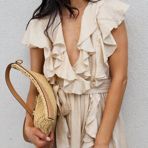 May include: A beige sleeveless dress featuring a deep V-neckline with cascading ruffles along the bodice and a matching waist tie. The dress is paired with a tan woven straw handbag that has a structured handle and a smooth leather trim.