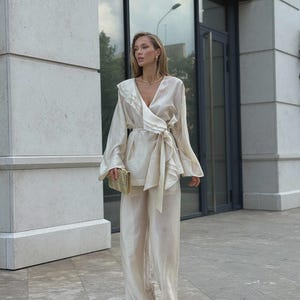 以下が含まれることがあります： クリーム色のツーピースセット。フリル付きのラペルとウエストで結ぶリボンが特徴の長袖ラップトップと、お揃いのワイドレッグパンツで構成されています。生地にはほのかな光沢があり、小さなクラッチバッグを合わせています。