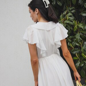 May include: A white dress featuring a wide collar, flutter sleeves, and a gathered waistline. The garment is paired with a light-colored rectangular hair claw clip. The dress has a textured fabric finish and a flowing skirt that falls below the waist.