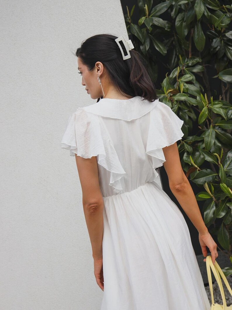 May include: A white dress featuring a wide collar, flutter sleeves, and a gathered waistline. The garment is paired with a light-colored rectangular hair claw clip. The dress has a textured fabric finish and a flowing skirt that falls below the waist.