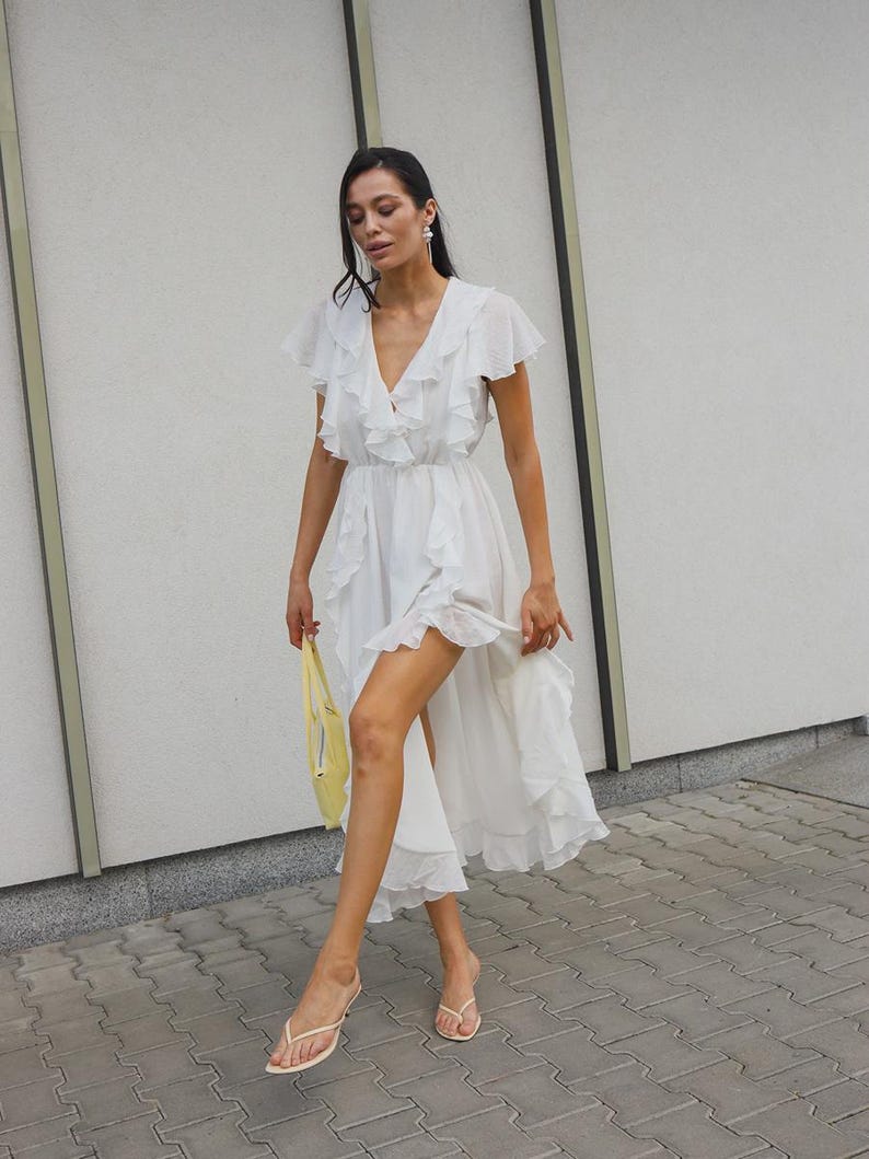 May include: A white midi dress featuring a deep V-neckline, flutter sleeves, and cascading ruffles that run along the front and hemline. The dress has a high-low silhouette and is paired with a small yellow handbag and light-colored thong sandals.