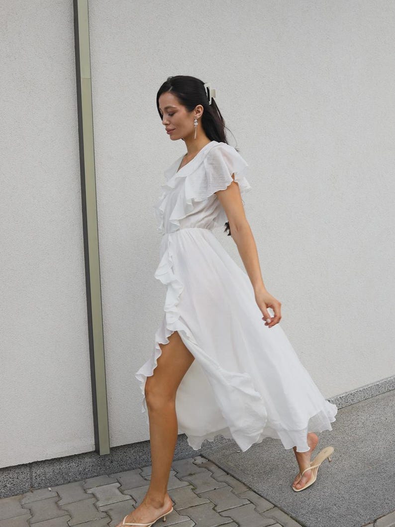 May include: A white midi dress featuring a V-neckline, short flutter sleeves, and a cascading ruffle detail that runs along the front opening. The lightweight fabric creates a flowing silhouette with a high side slit, paired with beige heeled sandals.