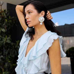 May include: A light blue sleeveless dress featuring a deep V-neckline with cascading ruffles along the bodice. The garment is paired with large, circular silver-toned earrings that have a textured finish and a central white pearl accent.