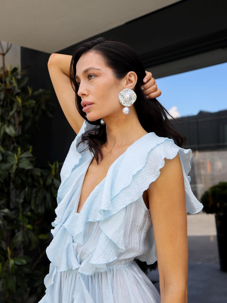 May include: A light blue sleeveless dress featuring a deep V-neckline with cascading ruffles along the bodice. The garment is paired with large, circular silver-toned earrings that have a textured finish and a central white pearl accent.