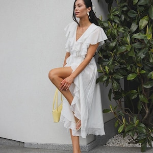 May include: A white midi dress featuring a V-neckline, short ruffled sleeves, and a tiered ruffled skirt with a high-low hemline. The dress is paired with a small yellow handbag and beige thong-style heeled sandals with thin straps.