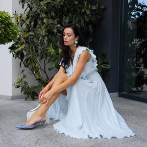 May include: A light blue sleeveless maxi dress featuring a V-neckline, ruffled shoulder details, and a flowing skirt with a textured fabric finish. The dress is paired with pointed-toe slingback heels in a matching light blue shade.