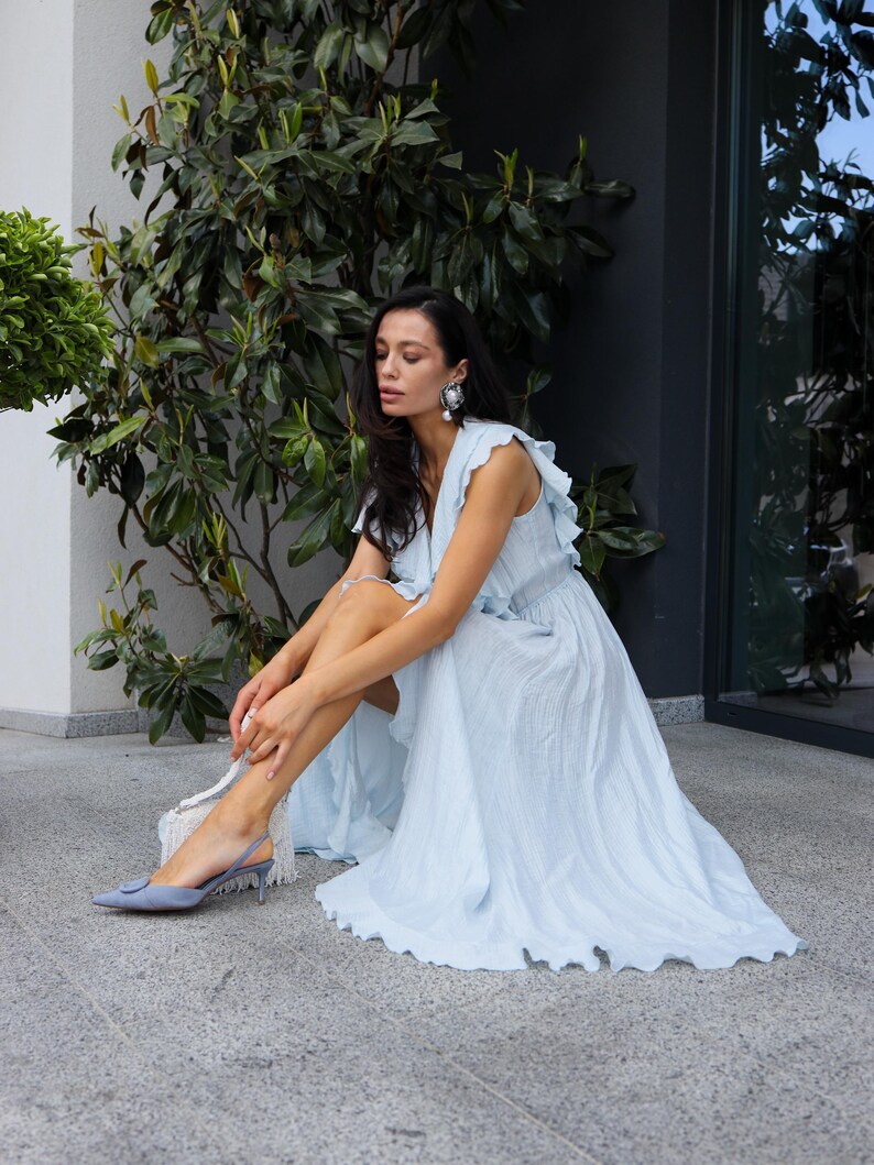 May include: A light blue sleeveless maxi dress featuring a V-neckline, ruffled shoulder details, and a flowing skirt with a textured fabric finish. The dress is paired with pointed-toe slingback heels in a matching light blue shade.