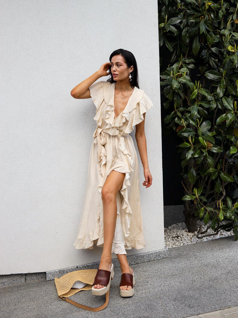 May include: A beige midi-length wrap dress featuring a deep V-neckline, short flutter sleeves, and cascading ruffles along the front opening. The dress includes a matching fabric tie at the waist and is paired with brown leather platform slides.