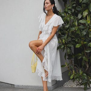 May include: A white midi dress featuring a V-neckline, short ruffled sleeves, and a tiered ruffled skirt with a high-low hemline. The dress is paired with a small yellow handbag and beige thong-style heeled sandals with thin straps.