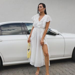 May include: A white midi dress featuring a V-neckline, flutter sleeves, and cascading ruffles along the front and skirt. The dress has a cinched waist and a high side slit. A small yellow handbag and beige thong sandals complete the ensemble.
