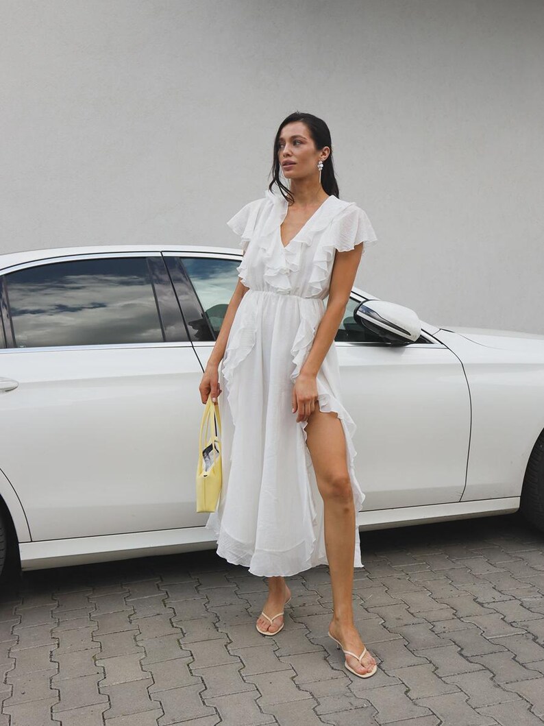 May include: A white midi dress featuring a V-neckline, flutter sleeves, and cascading ruffles along the front and skirt. The dress has a cinched waist and a high side slit. A small yellow handbag and beige thong sandals complete the ensemble.