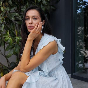 May include: A light blue sleeveless dress featuring a V-neckline, a fitted waist, and cascading ruffles along the shoulders and bodice. The fabric has a subtle, fine-textured finish, and the garment flows into a full, loose-fitting skirt.