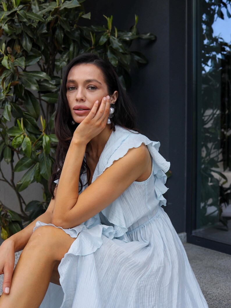 May include: A light blue sleeveless dress featuring a V-neckline, a fitted waist, and cascading ruffles along the shoulders and bodice. The fabric has a subtle, fine-textured finish, and the garment flows into a full, loose-fitting skirt.