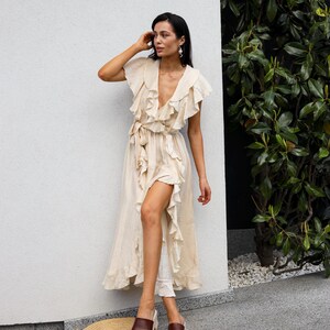 May include: A beige midi-length wrap dress featuring a deep V-neckline, short flutter sleeves, and cascading ruffles along the front opening. The dress includes a matching fabric tie at the waist and is paired with brown leather platform slides.