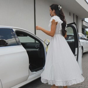 May include: A white midi dress featuring ruffled sleeves, a cinched waist, and a ruffled hemline. The dress is paired with beige thong-style kitten heel sandals and a white hair claw clip, shown from a side profile view near a white car.
