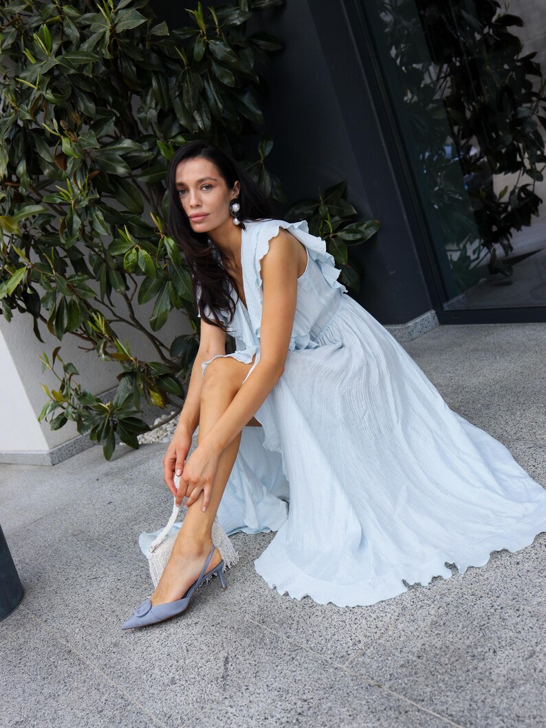 May include: A light blue sleeveless maxi dress with a ruffled neckline and a textured fabric finish. The dress features a flowing skirt with a high-low hemline. Paired with light blue pointed-toe slingback heels with a decorative buckle detail.