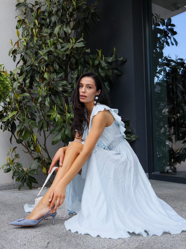 May include: A light blue sleeveless midi dress with a ruffled neckline and a textured, crinkled fabric finish. The dress features a flowing, A-line skirt that reaches mid-calf, paired with matching light blue pointed-toe slingback heels.