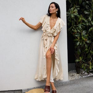 May include: A beige midi-length wrap dress featuring a deep V-neckline, short flutter sleeves, and cascading ruffle trim along the front opening. The dress includes a matching fabric tie at the waist and is paired with brown platform slide sandals.