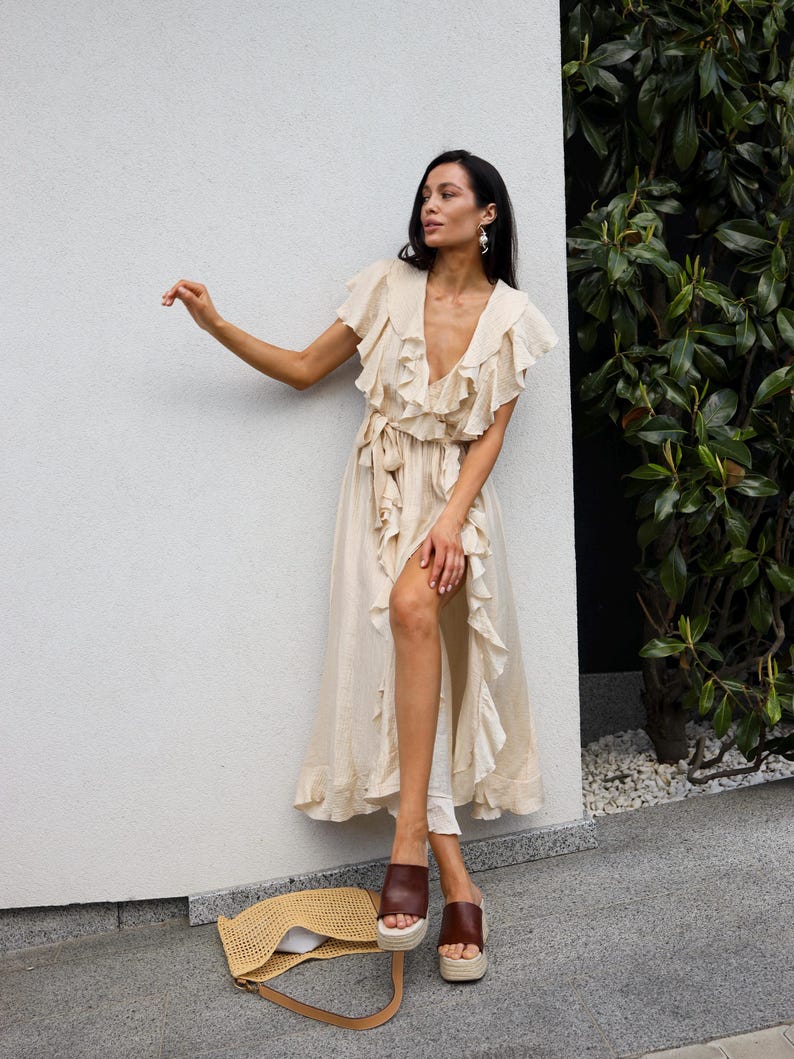 May include: A beige midi-length wrap dress featuring a deep V-neckline, short flutter sleeves, and cascading ruffle trim along the front opening. The dress includes a matching fabric tie at the waist and is paired with brown platform slide sandals.