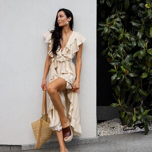 May include: A cream-colored wrap dress featuring a deep V-neckline, ruffled sleeves, and a ruffled hemline. The dress includes a matching fabric tie at the waist. A tan woven straw tote bag with leather handles and brown platform slide sandals.