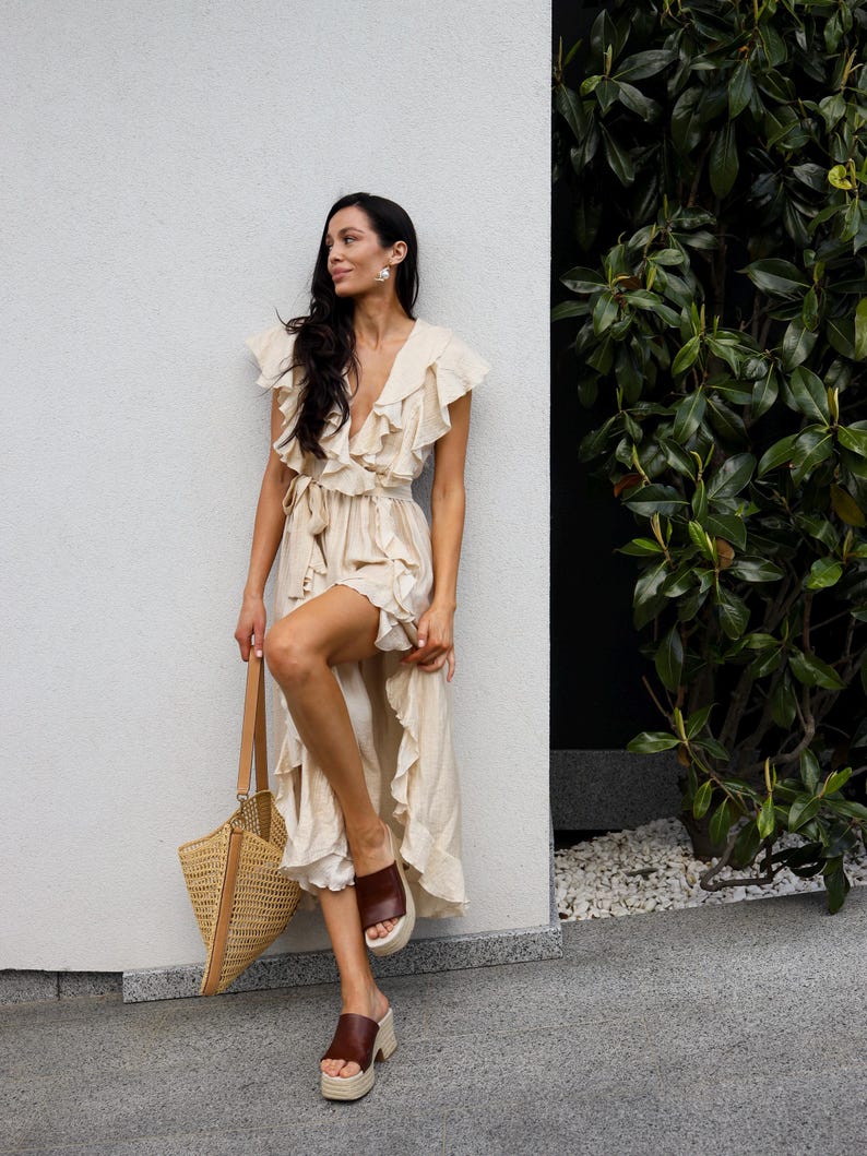 May include: A cream-colored wrap dress featuring a deep V-neckline, ruffled sleeves, and a ruffled hemline. The dress includes a matching fabric tie at the waist. A tan woven straw tote bag with leather handles and brown platform slide sandals.