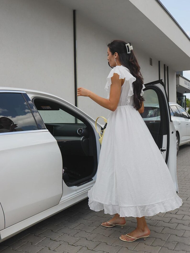 May include: A white midi dress featuring ruffled sleeves, a cinched waist, and a ruffled hemline. The dress is paired with beige thong-style kitten heel sandals and a white hair claw clip, shown from a side profile view near a white car.
