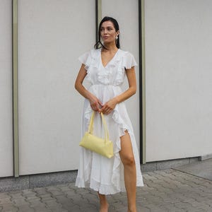 May include: A white midi dress featuring a V-neckline, short flutter sleeves, and cascading ruffles along the front and side slit. The dress is paired with a pale yellow rectangular shoulder bag and white thong-style heeled sandals.