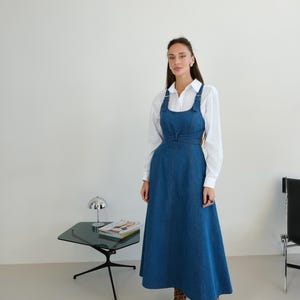 Puede incluir: Vestido peto de mezclilla azul con falda acampanada y tirantes ajustables. El vestido se lleva sobre una camisa blanca. La modelo lleva botas con estampado de leopardo. Una pequeña mesa negra con una lámpara y revistas está en el fondo.