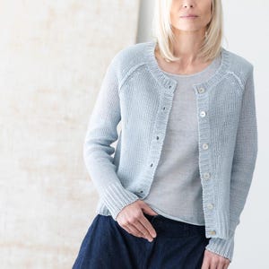 May include: A light blue cardigan with a round neckline and button-up front. The cardigan is made of a knitted material and is paired with a light gray top and dark blue denim pants. The outfit is simple and elegant.