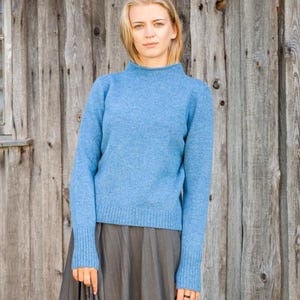 May include: A light blue sweater with a mock neck and long sleeves. The sweater is paired with a gray skirt. The background is a weathered wooden wall. The sweater's cuffs and hem are ribbed.