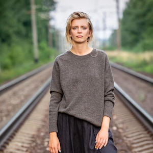 May include: A person wearing a gray crew neck sweater and black skirt stands on railroad tracks. The sweater has long sleeves and a relaxed fit. The background features green trees and a cloudy sky.