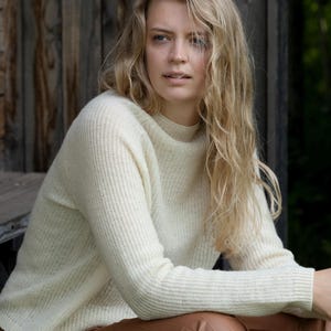 May include: A cream-coloured, ribbed knit jumper with a mock neck. The long-sleeved jumper is worn by a person with long, wavy blonde hair. The background is a wooden structure.