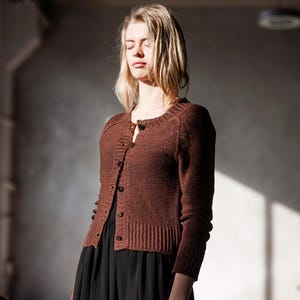 Puede incluir: Un cárdigan de punto marrón con botones en la parte delantera y mangas largas. El cárdigan se combina con un vestido negro. La modelo tiene el pelo rubio y está en una habitación con luz natural.