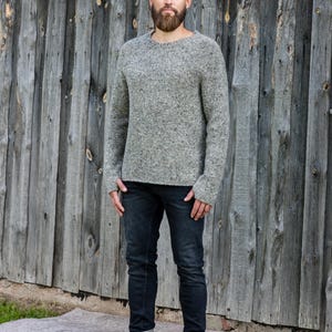 May include: A long-sleeved, crew-neck sweater in a speckled grey knit pattern, paired with dark charcoal denim jeans and grey lace-up ankle boots with a rounded toe and a thick rubber sole, worn by a person with a short beard and short hair.