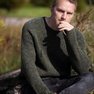 Peut inclure: Une personne porte un pull ras du cou vert foncé avec des mouchetures de couleur, associé à un jean gris foncé. Le pull a un tricot texturé, et la personne est assise à l'extérieur avec une expression pensive.