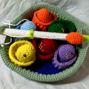 May include: A sage green crocheted basket containing five small, colorful crocheted creatures, each nestled in its own matching crocheted pod. A long, white and lime green crocheted fishing rod with an orange tip rests across the top of the basket.