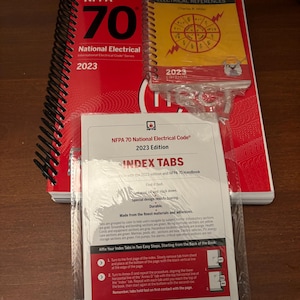 May include: A stack of electrical reference books and index tabs. The books include "NFPA 70 National Electrical Code 2023" and "Ugly's Electrical References 2023 Edition." Index tabs are included for easy navigation.