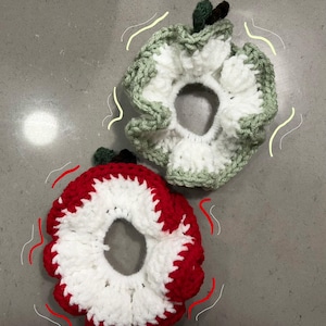May include: Two handmade crochet scrunchies in the shape of apples. One is red and white, the other is green and white. Each has a small green stem. These hair accessories add a playful touch to any hairstyle.