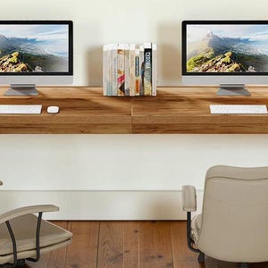 May include: A wooden desk with two computer monitors, two desk lamps, and two chairs. The desk has a natural wood finish and is long enough for two people to work comfortably. The chairs are beige and have armrests.