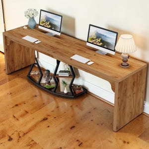 May include: A long, light brown wood-grain desk featuring two computer monitors, two white keyboards, and a white lamp. The desk is supported by a black metal frame with integrated shelving units holding small decorative items and books.